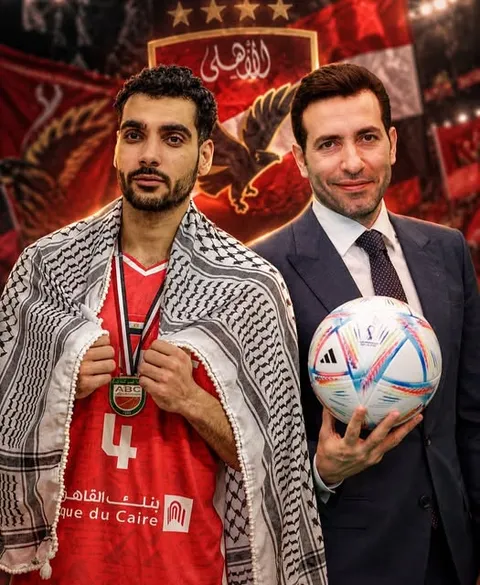 رجلان متجاوران؛ أحدهما بقميص رياضي أحمر ووشاح وميدالية، والآخر ببدلة يحمل كرة، أمام خلفية شعار الأهلي الأحمر.